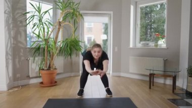 Kendinden emin, iri yarı beyaz bir kadının kameraya bakıp gülümsediği geniş açılı bir portresi. Kilolu, genç ve güzel bir bayan sabah evde antrenman yapıyor. Fitness ve kendini geliştirme.