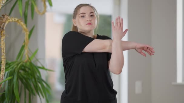 Sérieux concentré sportive en surpoids échauffer les muscles de la main de formation à la maison le matin. Portrait médian de la jeune femme caucasienne concentrée de plus de taille travaillant à l'intérieur. — Video