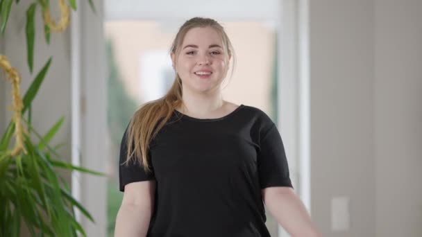 Inquadratura di mezzo di bella sportiva positiva plus-size utilizzando bottiglie d'acqua come manubri sorridenti e guardando la fotocamera. Ritratto di donna caucasica sicura di sé che si esercita a casa. — Video Stock