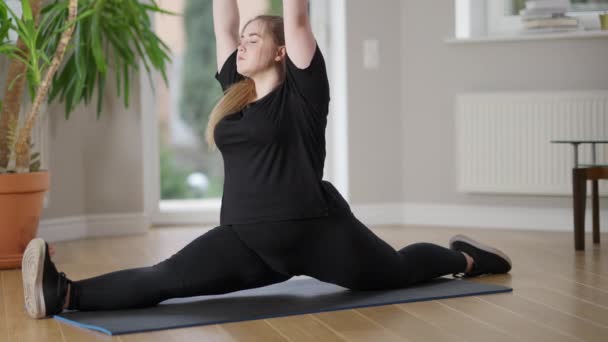 Vue de côté plan large de charmante jeune femme obèse assise en ficelle pose sur le tapis d'exercice faire de l'exercice respiratoire et rire. Sportive caucasienne positive s'entraînant à l'intérieur à la maison. Concept sportif. — Video