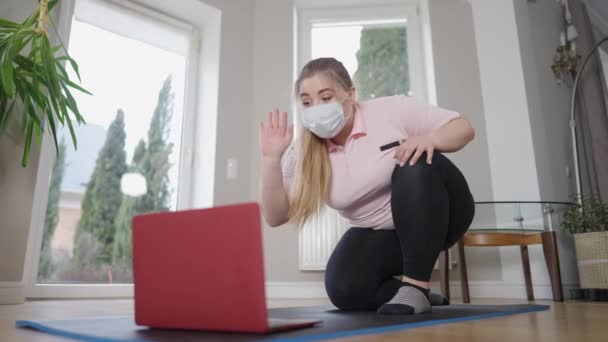 Portrait of positive beautiful obese woman in Covid-19 face mask waving closing laptop and leaving. Happy overweight Caucasian sportswoman saying goodbye after online training at home on pandemic. — Stock Video