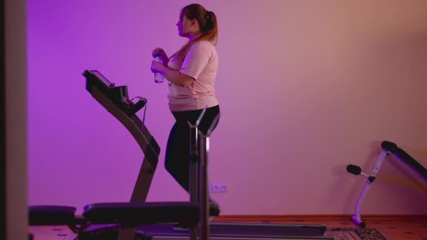 Vista laterale della paffuta donna caucasica che cammina sul tapis roulant bevendo acqua rinfrescante. Ampio colpo di persistente motivato giovane sportiva formazione in palestra a casa al chiuso. Concetto di sport e resistenza. — Video Stock