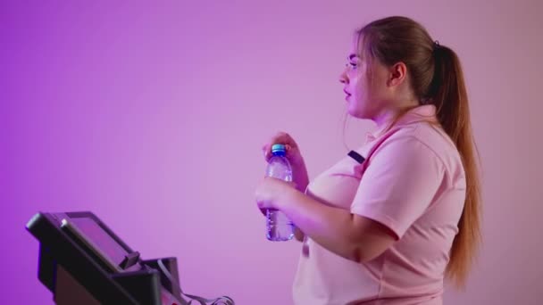 Vue latérale de la transpiration jeune femme en surpoids marchant sur tapis roulant boire de l'eau rafraîchissante. Plan du milieu de motivé Caucasien plus-taille sportive formation à l'intérieur. — Video