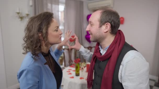 Adult couple kissing holding hands together in heart shape at background and smiling looking at camera. Positive loving happy Caucasian man and woman posing indoors on Valentines. — Stock Video