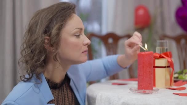 Seitenansicht der schönen konzentrierten Frau, die rote Kerze mit Streichholz anzündet. Lächelnde kaukasische Dame, die zu Hause eine romantische Valentinsfeier vorbereitet. Liebe und Urlaubskonzept. — Stockvideo