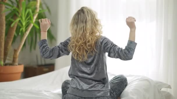 Vista posteriore di donna caucasica bionda magra in pigiama che si estende seduta sul letto a casa. Ampia ripresa del giovane millenario dai capelli ricci che si sveglia in camera da letto al chiuso godendo del mattino del fine settimana. — Video Stock