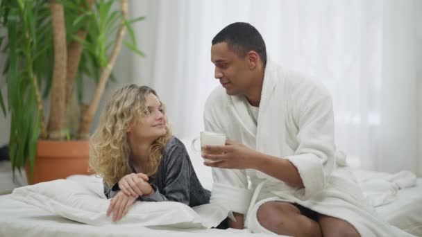 Afro-Amerikalı bir adam beyaz bir kadına sabah kahvesi getiriyor. Evdeki yatak odasında kız arkadaşına bakan mutlu bir erkek arkadaşın geniş açılı portresi. Aşk ve romantizm. — Stok video