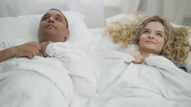 Νεαρό ευτυχισμένο ζευγάρι που κρύβεται κάτω από άσπρες κουβέρτες αγκαλιά ξαπλωμένο στο κρεβάτι. Χαμογελώντας Αφροαμερικανός και Καυκάσια γυναίκα διασκεδάζουν στην κρεβατοκάμαρα το πρωί. Αγάπη και δέσιμο. — Αρχείο Βίντεο