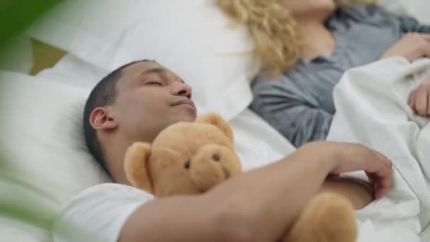 Közelkép afro-amerikai fiatalemberről, amint Teddy macit ölelget kényelmes ágyban, homályos, felismerhetetlen fehér nővel a háttérben. Boldog férj pihen feleséggel a hálószobában. — Stock videók