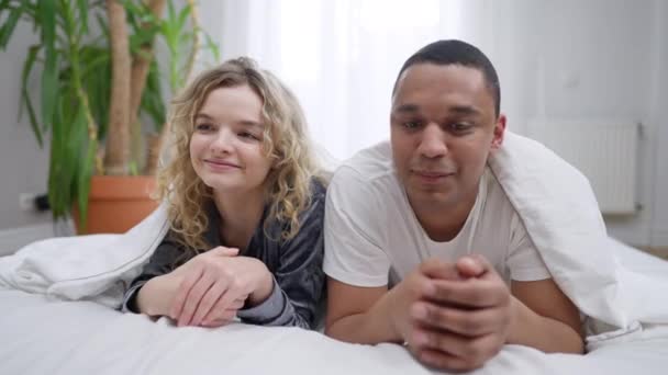 Verlegen millennial interraciale paar kijken naar elkaar glimlachend liggend in bed onder deken. Positieve gelukkige liefhebbers genieten van de ochtend in de slaapkamer. Afro-Amerikaanse man raakt neus van blanke vrouw. — Stockvideo