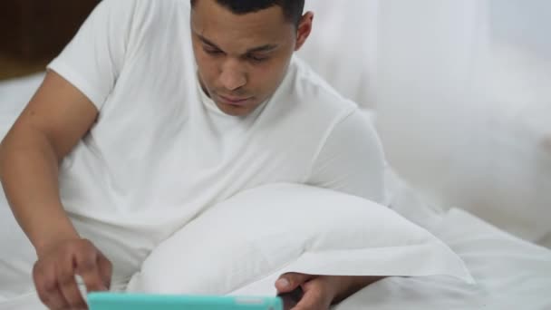 Absorbido joven afroamericano mensajería en línea en las redes sociales en la tableta acostado en la cómoda cama blanca sonriendo. Retrato de millennial relajado en el dormitorio por la mañana. — Vídeos de Stock