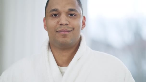 Retrato de cerca de un joven afroamericano feliz vestido con albornoz blanco mirando a la cámara sonriendo de pie en la ventana del dormitorio. Positivo relajado guapo milenario posando en casa por la mañana. — Vídeos de Stock