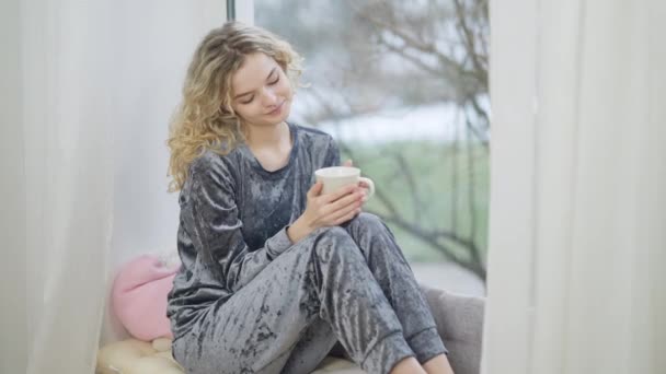 Encantadora mujer sonriente feliz sentada en el alféizar de la ventana sosteniendo la taza de café mirando por la ventana. Romántico joven caucásico milenario en pijama disfrutando de la mañana en interiores pensando. — Vídeos de Stock