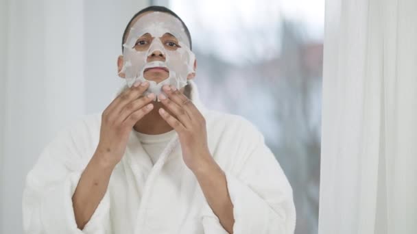 Spegel POV av ung afroamerikansk man i vit badrock sätta på renande ansiktsmask stående i sovrum på morgonen. Självsäker stilig kille som tar hand om ansiktet hemma. — Stockvideo