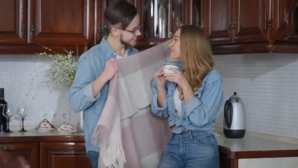 Ung smilende flot mand ind i køkkenet indpakning smuk kvinde i tæppe. Glædelig kærlig kærlig kæreste tager sig af charmerende kæreste om morgenen. Forholdet og enhed. – Stock-video