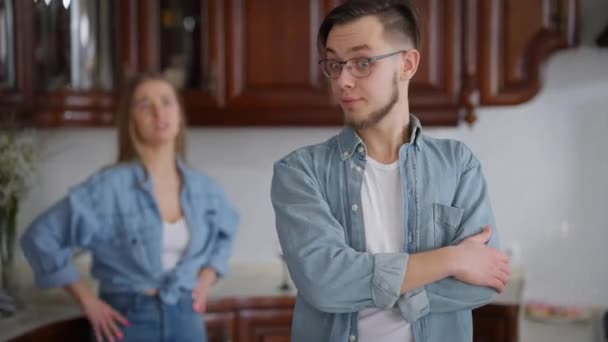 Junger gestresster Mann mit schockiertem Gesichtsausdruck und verschränkten Händen, der in der Küche steht, während eine wütende Frau gestikuliert. Porträt eines kaukasischen Freundes mit streitender Freundin im Haus. — Stockvideo