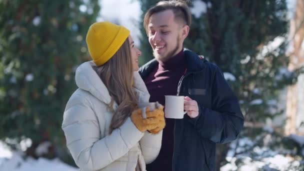 Medium shot portret van gelukkig jong stel buiten staan met koffiekopjes glimlachend praten. Blanke man en vrouw genieten van wintersportvakantie op zonnige dag. Verbindingsconcept. — Stockvideo