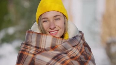 Sarı şapkalı, battaniyeye sarılı mutlu genç bir kadının dışarıda kameraya bakarken çekilmiş portresi. Kış günü yavaş çekimde poz veren neşeli, güzel, zayıf milenyumun yakın plan yüzü..