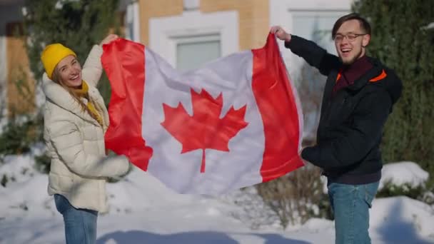 Jovens canadenses felizes posando com a bandeira nacional balançando em mãos ao ar livre no ensolarado quintal de inverno. Retrato de homem e mulher sorridentes e alegres olhando para a câmera à luz do sol. — Vídeo de Stock