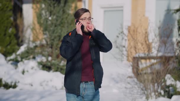 Młody biały mężczyzna rozmawia przez telefon stojąc na zewnątrz w słoneczny zimowy dzień. Średni portret pewnego siebie przystojnego tysiąclecia w okularach i ciepłych ubraniach ze smartfonem w świetle słonecznym. — Wideo stockowe