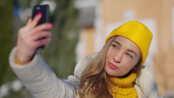 Encantadora joven mujer milenaria tomando selfie en el teléfono inteligente de pie en el soleado complejo de invierno al aire libre. Primer plano retrato de alegre y relajada dama sonriente viajando. Turismo y adicción a los dispositivos. — Vídeo de stock