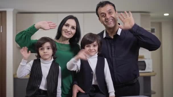 Happy cheerful multiethnic family waving smiling looking at camera indoors. Portrait of joyful Caucasian wife Middle Eastern husband and twin brothers posing at home in living room. — Wideo stockowe