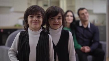 Orta boy Orta Doğulu ikiz kardeşler oturma odasında poz veriyor ve arka planda iki ebeveyn oturuyor. Gülümseyen çocuklar kameraya bakıyorlar..