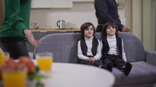 Middle Eastern twin brothers sitting on couch watching TV as couple of interracial parents entering living room hugging sons. Happy family on weekend at home indoors. Leisure and unity concept. — Wideo stockowe