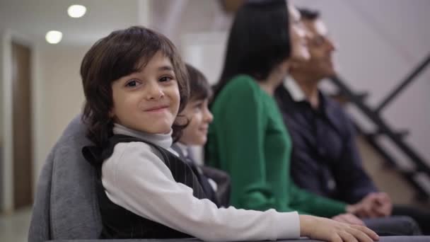 Happy cute boy looking at camera smiling with positive family resting at background watching TV sitting on cozy couch. Portrait of cheerful Middle Eastern child posing in living room indoors. — Stock Video