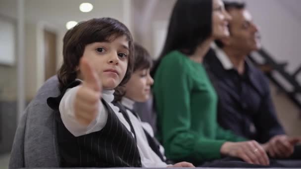 Bonito menino do Oriente Médio positivo mostrando polegar com irmão gêmeo e pais assistindo TV no fundo. Retrato de criança confiante posando em casa com a família se divertindo no fim de semana. — Vídeo de Stock