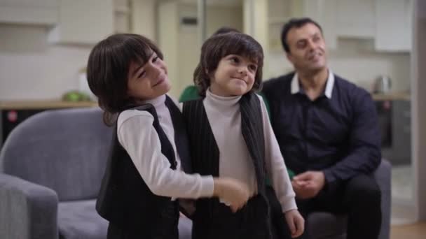Zwillingsbrüder umarmen sich lächelnd und blicken in die Kamera, während verschwommene Eltern im Hintergrund fernsehen. Positive Nahost-Kinder posieren zu Hause. — Stockvideo