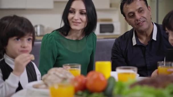 Homem e mulher sorridentes conversando com gêmeos bonitos jantando à noite. Mãe caucasiana positiva e pai do Oriente Médio conversando com filhos em casa no almoço. — Vídeo de Stock