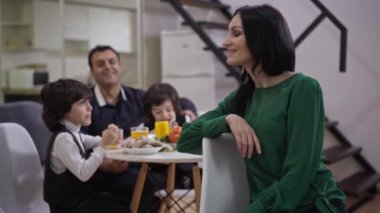 Oturma odasında neşeli Orta Doğulu erkek ve oğlanların arka planda yemek yediği mutlu beyaz kadın portresi. Olumlu gülümseyen anne karısı kameraya bakıyor..