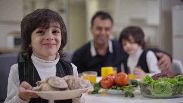 Portrait of cute boy holding plate with sweet delicious bakery looking at camera as blurred family enjoying dinner at background. Happy smiling Middle Eastern child posing indoors. — Stock Video