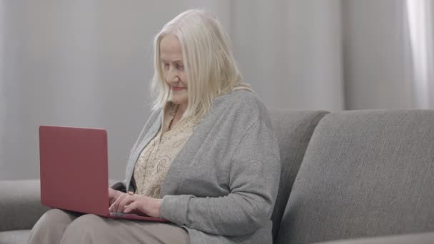 Боковой вид старой белой женщины с седыми волосами, печатающей на клавиатуре ноутбука, сидящей на уютном диване в гостиной. Современная бабушка переписывается дома. Современные технологии и стиль жизни. — стоковое видео