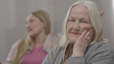 Mutsuz, gri saçlı, arka planda tartışan bulanık bir kadın olarak kameraya bakan mutsuz, yaşlı, beyaz bir kadının yakın plan portresi. Evde tatminsiz bir kızı olan üzgün yaşlı bir anne..