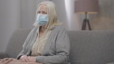 Coronavirus maskeli yaşlı gri saçlı kadının orta boy portresi kanepeye oturmuş kameraya bakıyor. Evde, Covid-19 pandemik tecritte yalnız yaşlı bir kadının portresi. Kilitleme kavramı.