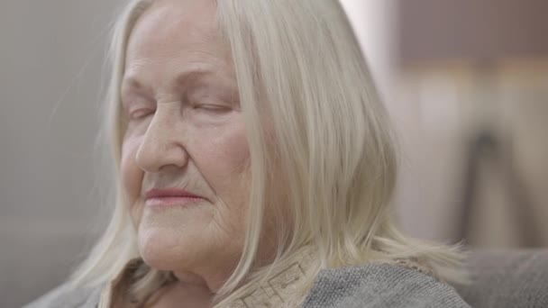 Retrato en la cabeza de una vieja mujer caucásica triste y solitaria con el pelo gris mirando a la cámara con expresión facial alterada. Desesperado frustrado jubilado solo en casa en la sala de estar en el interior. — Vídeo de stock