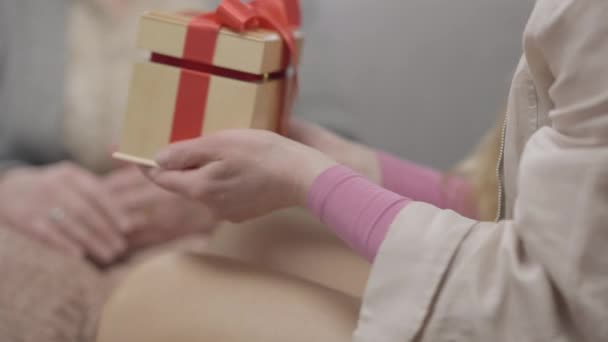 Camera follows gift box in female hands as woman giving present to old blurred Caucasian retiree. Friend congratulating pensioner on holiday indoors. Friendship and aging concept. — Stock Video