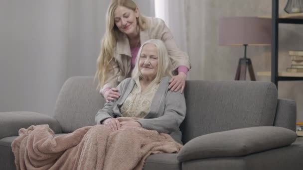 Широко снятый портрет улыбающейся счастливой старухи, сидящей на диване, как пожилая леди, обнимающая мать, смотрящая в камеру. Уверенная, расслабленная кавказская пенсионерка наслаждается выходными с дочерью дома позирующей. — стоковое видео
