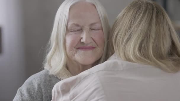 A boldog öreg kaukázusi anya portréja, ahogy otthon ölelgeti a lányát, miközben a kamerába néz. Közelkép mosolygós nyugodt, magabiztos kaukázusi öregasszonyról, aki pózol. Az öregedés és gondozás fogalma. — Stock videók