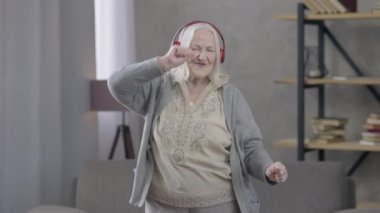 Rahatlamış, kulaklıklı, kulaklıkla dans eden, gülümseyen bir emekli. Evdeki hobisinden zevk alan beyaz saçlı, neşeli, yaşlı bir kadının orta boy portresi. Neşe ve mutluluk.