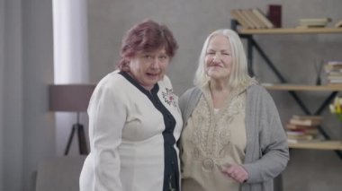 Oturma odasında dans eden iki aktif son sınıf öğrencisi kameraya bakıyor. Pozitif kadın emeklilerin hafta sonları evde buluşarak eğlendikleri bir portre. Neşe ve emeklilik yaşam tarzı.