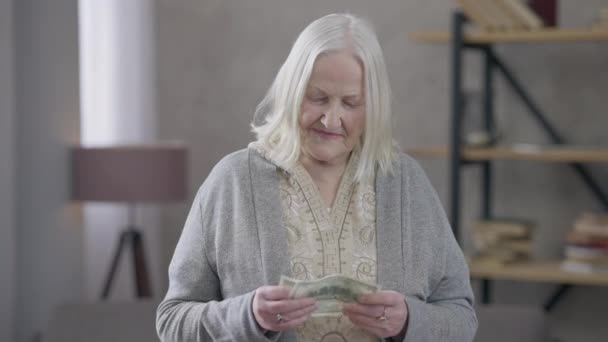 Gråhårig gammal kaukasisk kvinna som räknar suckande pengar med kameran. Stackars kvinna som poserar i vardagsrummet och har kontanter. Fattigdom och pensionering. — Stockvideo