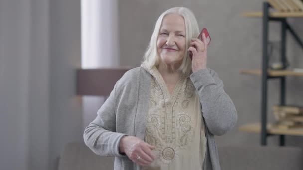 A boldog öreg fehér nyugdíjas arcképe, aki felveszi a telefont és mosolyog. Vidám idős nő, aki otthon áll a nappaliban. Boldogság és kommunikáció koncepciója. — Stock videók