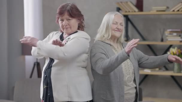 Magabiztos, idős, fehér nők keresztezik egymás kezét, miközben a kamerát nézik, ahogy háttal állnak egymásnak. Pozitív örömteli nyugdíjasok portréja a nappaliban. Magabiztos és öregedő. — Stock videók