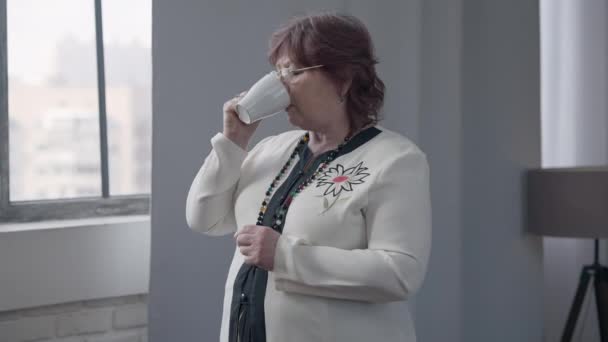 Magányos vörös hajú nő szemüvegben reggeli teát iszik, vagy kávét bámul ki az ablakon gondolkodva. Közepesen feldúlt kaukázusi nyugdíjas portréja otthon, egyedül.. — Stock videók
