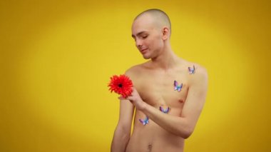 Gövdesinde kelebekler olan ve elinde kırmızı çiçeğe hayran olan abartılı, tuhaf bir adam. Beyaz erkek LGBTQI 'nin sarı arka planda poz verdiği orta boy bir fotoğraf..