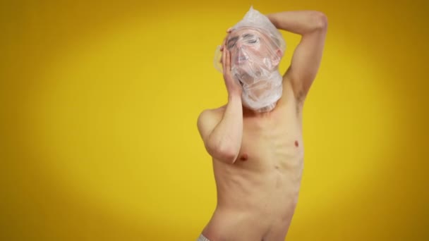 Portret van een jonge blanke man met plastic op het hoofd poserend op een gele achtergrond. Man Kaukasische homo verstikkend van sociale intolerantie concept. — Stockvideo