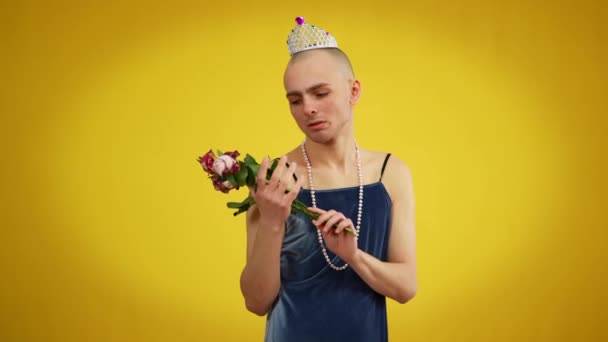 Jonge trieste homo in jurk en tiara met boeket verwelkte bloemen die naar de camera kijken. Portret van blanke mannelijke transgender poserend op gele achtergrond. — Stockvideo